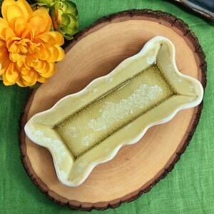 May include: A small, rectangular, ceramic dish with a wavy edge. The dish is glazed in a light green color with a darker green glaze inside. The dish is sitting on a wooden slice.