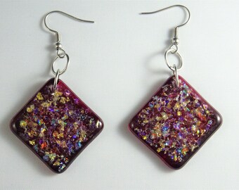 May include: A pair of earrings with a square-shaped glass pendant. The glass is a deep red color with iridescent gold and purple flecks. The earrings are attached to silver hooks.