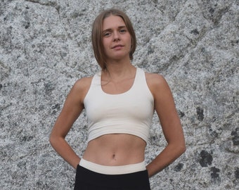 Può includere: Una donna indossa un top bianco corto e pantaloni neri con una fascia bianca in vita. È in piedi davanti a una grande roccia.