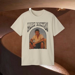 Puede incluir: Una camiseta beige con un estampado gráfico negro de un hombre sentado en una duna de arena. El gráfico incluye el texto "HARRIS DICKINSON" en una fuente estilizada.