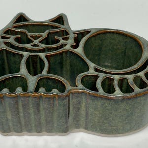 May include: A ceramic cat-shaped organizer in shades of green and brown. The cat design features cut-out details for the face, body, and paws, with multiple compartments for storage. The glaze has a glossy finish.