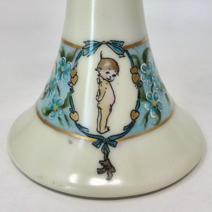 May include: A white ceramic pedestal with a blue and gold floral design and a painted image of a cherub. The cherub is surrounded by a wreath of forget-me-not flowers and hearts.