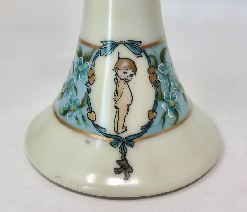 May include: A white ceramic pedestal with a blue and gold floral design and a painted image of a cherub. The cherub is surrounded by a wreath of forget-me-not flowers and hearts.