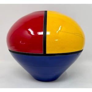 May include: A decorative ceramic vase with a geometric design. The top half is divided into red and yellow sections, separated by a black line. The bottom half is blue. The vase has a rounded top and a tapered base.