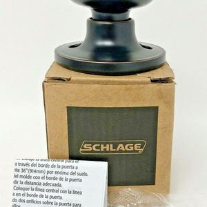 May include: A dark bronze door knob with a rounded handle and a circular base, sitting on a cardboard box. The box has the brand name "SCHLAGE" printed on it. Includes screws and instructions.