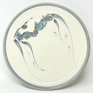 May include: A round ceramic plate with a light cream-colored surface and a gray rim. The plate features a delicate, abstract design in shades of blue, gray, and red. The design appears to be a stylized floral or organic motif.