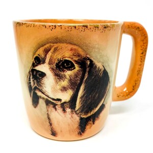 May include: A ceramic mug with a detailed illustration of a beagle's face. The mug has a peach-colored base with a matching handle and rim. The beagle's fur is depicted in shades of brown, black, and white, with a focused expression. The mug is designed for beverages.