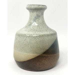 May include: A small, ceramic vase with a rounded body and a narrow neck. The vase features a color-blocked design with shades of brown, beige, and gray. The surface has a speckled texture, adding visual interest to the piece.