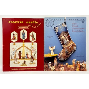 May include: Two vintage cross-stitch pattern books. One is red with the text "Creative Needle" and "Away in a Manger." The other is blue with a Christmas stocking design and the text "The First Christmas Stocking."