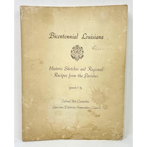 May include: A vintage cookbook titled "Bicentennial Louisiana" with the subtitle "Historic Sketches and Regional Recipes from the Parishes." The cover is a faded tan color with dark brown text and a decorative emblem. Sponsored by the Cultural Arts Committee.