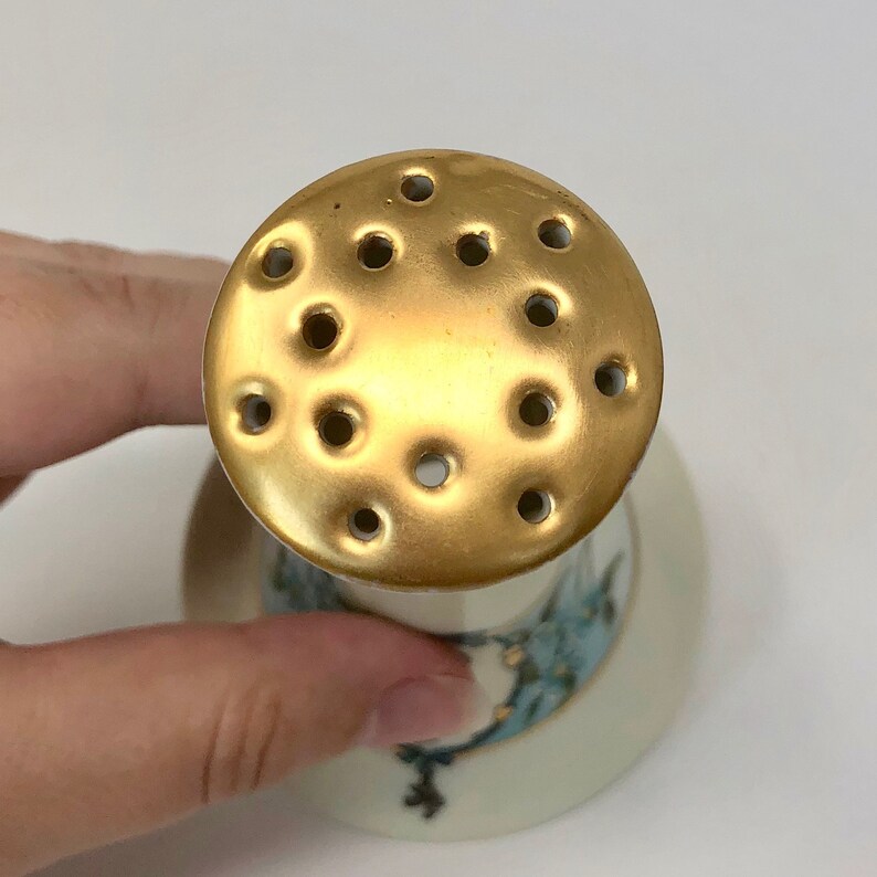 May include: A white ceramic salt shaker with a gold-colored top. The top has a series of holes for dispensing salt.