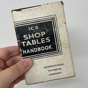 May include: Vintage "ICS Shop Tables Handbook" with black text on a cream-colored cover. The handbook is held by a hand and has the text "International Textbook Company" at the bottom. The cover has a black border.