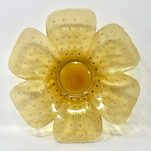 May include: A flower-shaped glass bowl in a translucent amber color. The bowl features six rounded petals with a textured surface pattern consisting of small, evenly spaced rectangular indentations radiating from a smooth, circular center.