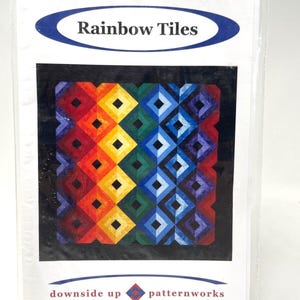 May include: A package featuring a colorful quilt pattern titled "Rainbow Tiles." The design showcases a geometric pattern of diamonds in a rainbow of colors, including red, orange, yellow, green, blue, and purple. The package also includes the text "downside up patternworks."