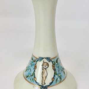 May include: A white porcelain flower frog with a gold rim and a blue and gold floral design featuring a cherub. The frog has a round base and a tall, slender neck with holes for flower stems.
