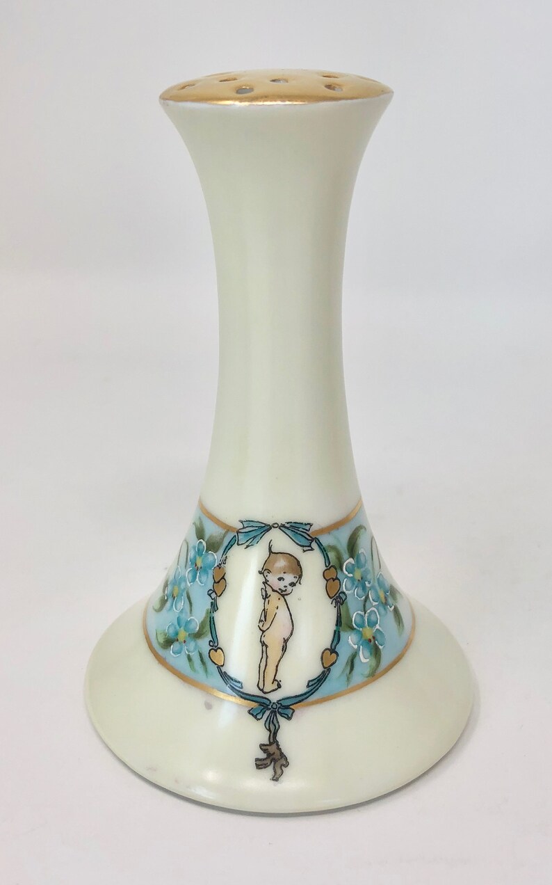 May include: A white porcelain flower frog with a gold rim and a blue and gold floral design featuring a cherub. The frog has a round base and a tall, slender neck with holes for flower stems.