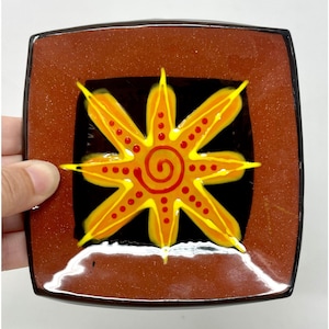 May include: Square ceramic plate with a brown border and a black center. The center features a yellow and orange sunburst design with a spiral center and red dots. The plate is held by a hand.