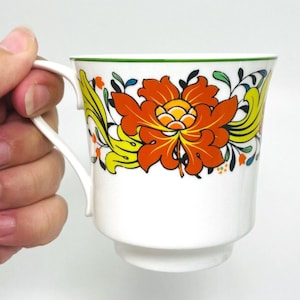 May include: White ceramic mug with a large orange and yellow floral design. The mug has a green rim and a curved handle. The floral design includes green, blue, and orange accents. The mug is held by a hand.