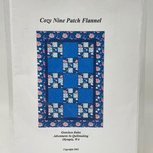 May include: A package featuring a quilt pattern titled "Cozy Nine Patch Flannel." The pattern design includes nine blue squares with floral and patterned accents. The package also includes the author's name, company, and copyright information.