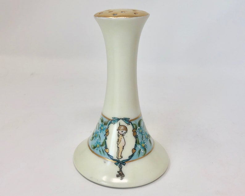 May include: White porcelain salt shaker with a blue and gold floral design and a small child figure in the center. The top of the shaker has a gold rim and holes for dispensing salt.