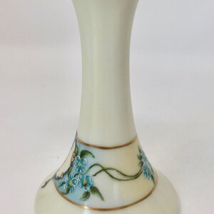 May include: A white porcelain vase with a gold rim and blue forget-me-not flowers painted on the side. The vase has a flared base and a narrow neck with holes for flower stems.