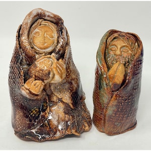 May include: Two ceramic sculptures of figures wrapped in textured cloaks. The figures have detailed faces and are glazed in shades of brown, tan, and green. One figure holds a bundle, while the other holds a leaf-like object.