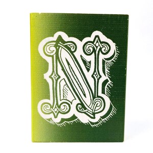 May include: A wooden block with a gradient green and yellow background. A white, ornate letter "N" is centered on the block. The letter has a decorative, vintage style with swirls and flourishes.