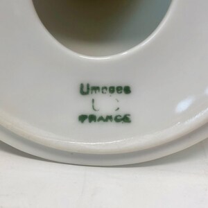 May include: Close-up of the bottom of a white ceramic object with a green mark that reads "Umcoee" and "France".
