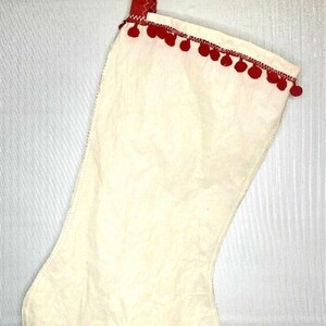 May include: Cream-colored Christmas stocking with red trim and pom-poms. The stocking has a red ribbon loop with small gold bells and a red ribbon. The top edge is decorated with red pom-poms.