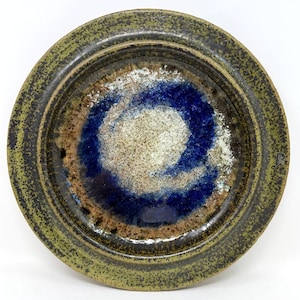 May include: A handmade ceramic plate with a unique design. The plate features a central swirl of blue and white, surrounded by brown and a textured green rim. The plate is round and has a rustic, artisanal feel.