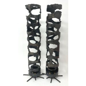 May include: Two dark metal candle holders with a textured, open design. Each holder is cylindrical with irregular cutouts and sits on a star-shaped base. The candle holders are a unique decorative item.