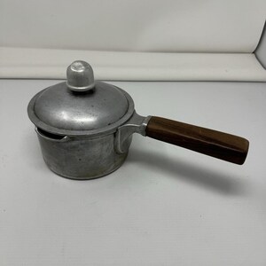 May include: A small aluminum saucepan with a matching lid and a dark brown wooden handle. The pot features a rounded body with a small pouring spout on the side. The lid has a central knob handle, and the overall finish of the metal is matte.
