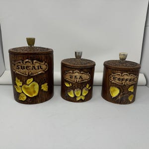 Könnte beinhalten: Set aus drei braunen Keramikbehältern mit Deckeln. Die Behälter sind mit "SUGAR", "TEA" und "COFFEE" in goldfarbenen Buchstaben beschriftet. Jeder Behälter hat ein strukturiertes Holzmaserungsdesign und gelbe Fruchtdetails. Die Deckel haben kleine Knöpfe.