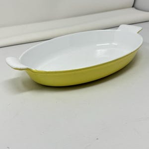 May include: Oval-shaped baking dish with a white interior and a yellow exterior. The dish has two small handles and is made of ceramic. The dish is empty and ready to be used for baking or serving.