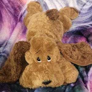 May include: A plush brown dog toy with large floppy ears and a dark brown spot on its head. The dog has large, sad-looking eyes and a black nose. It is lying on a colorful, patterned blanket.