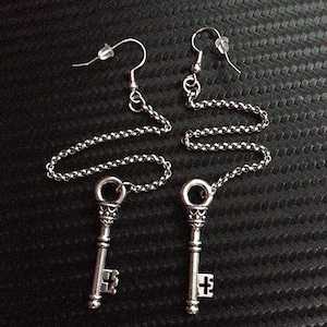 May include: A pair of silver key earrings with a chain extending from the key. The keys have ornate details and a cross-shaped design at the bottom.