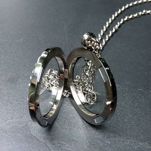 May include: Open silver locket pendant on a silver chain. The locket is oval-shaped and opens to reveal a small chain inside. The locket is displayed against a dark background.