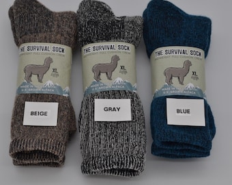 May include: Three pairs of alpaca wool socks in beige, gray, and blue. Each sock is labeled "The Survival Sock" with an alpaca illustration and the text "Made in the USA with U.S. Grown Alpaca".