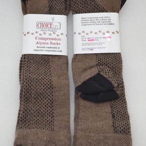May include: A pair of brown and black compression socks with a textured knit pattern. Each sock features a white paper band with the text Choice Alpaca Products, Compression Alpaca Socks, and details about fibre content, sizing, and care instructions.