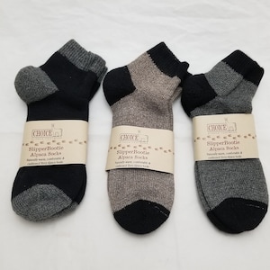 May include: Three pairs of alpaca slipper socks in black, brown, and gray. The socks are made of a soft, comfortable terry alpaca fabric. The packaging says "Choice Alpaca Footwear" and "Slipper Bootie Alpaca Socks".