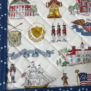 May include: Quilted fabric with a patriotic theme. The design features historical buildings, the Liberty Bell, soldiers, a ship, and the text "UNITED WE STAND" and "76". The border is navy blue with white stars.