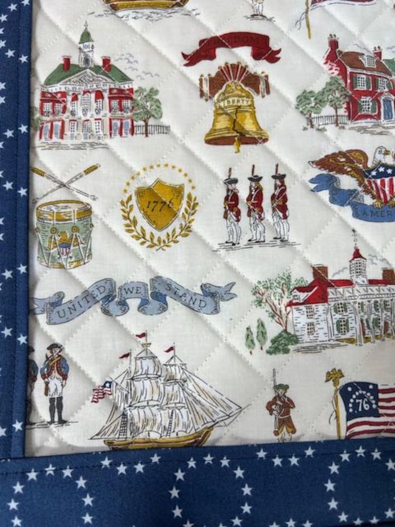 May include: Quilted fabric with a patriotic theme. The design features historical buildings, the Liberty Bell, soldiers, a ship, and the text "UNITED WE STAND" and "76". The border is navy blue with white stars.