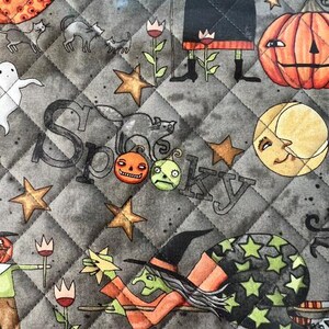 May include: A quilted fabric featuring a gray background with a diamond pattern. The print displays witches, black cats, pumpkins, ghosts, stars, and a crescent moon. The word Spooky is written in a stylized font among the illustrations.