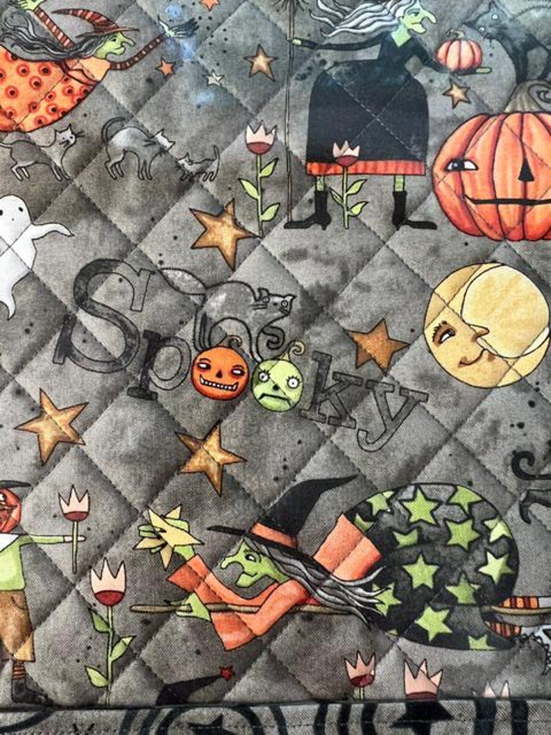 May include: A quilted fabric featuring a gray background with a diamond pattern. The print displays witches, black cats, pumpkins, ghosts, stars, and a crescent moon. The word Spooky is written in a stylized font among the illustrations.