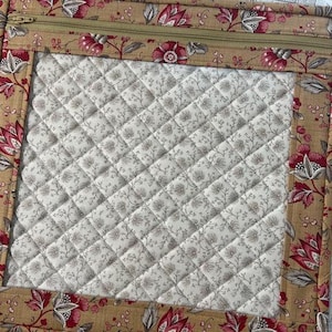 May include: A quilted project bag with a floral print border in shades of tan, red, and green. The bag features a clear window and a zipper closure. The interior is a quilted pattern in a neutral color.