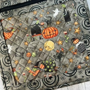 May include: A quilted fabric pouch featuring a Halloween-themed print with witches, pumpkins, ghosts, black cats, and stars on a gray background. The pouch has a black zipper closure and a border pattern of black circles and starbursts.