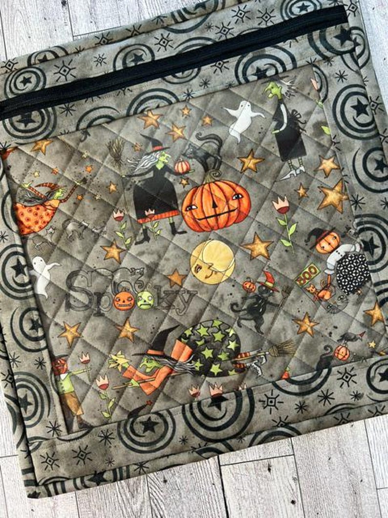 May include: A quilted fabric pouch featuring a Halloween-themed print with witches, pumpkins, ghosts, black cats, and stars on a gray background. The pouch has a black zipper closure and a border pattern of black circles and starbursts.