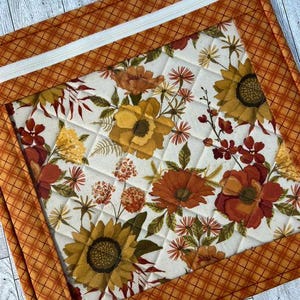 May include: A square quilted pouch featuring a central panel with a cream background and a floral print of sunflowers, daisies, and autumn-toned blossoms. The pouch is framed by an orange and brown plaid border and includes a white zipper closure.