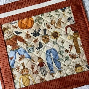 May include: A quilted fabric pouch featuring a cream-colored center panel with a repeating autumn print of scarecrows, pumpkins, sunflowers, squirrels, and black birds. The pouch is framed by a solid burnt orange border and a white zipper.