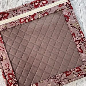 Puede incluir: Una bolsa rectangular acolchada con un borde de estampado floral y una cremallera de color claro. El interior es de color malva apagado liso con un patrón de diamantes. El borde floral presenta flores rojas y blancas sobre un fondo malva.